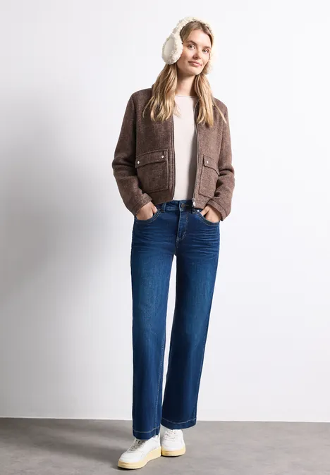 Een lachende vrouw draagt een bruine jas, witte trui, blauwe wijde jeans en witte sneakers voor een witte achtergrond.