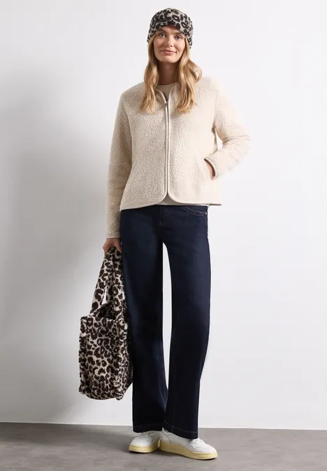 Bandeau en peluche à motif léopard auburn brown Frau mit beiger Teddyjacke, schwarzer Mütze, dunklen Jeans, weißen Sneakern und Leopardenmuster-Tasche vor weißer Wand.