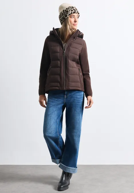 Frau in brauner Steppjacke, weiten Jeans, schwarzen Stiefeln und gemusterter Wintermütze vor weißem Hintergrund.