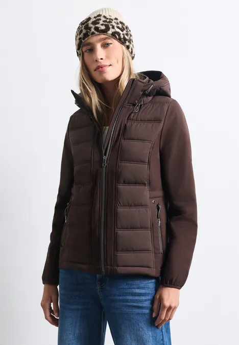 Frau in brauner Steppjacke, Jeans und Leopardenmütze vor hellem Hintergrund.