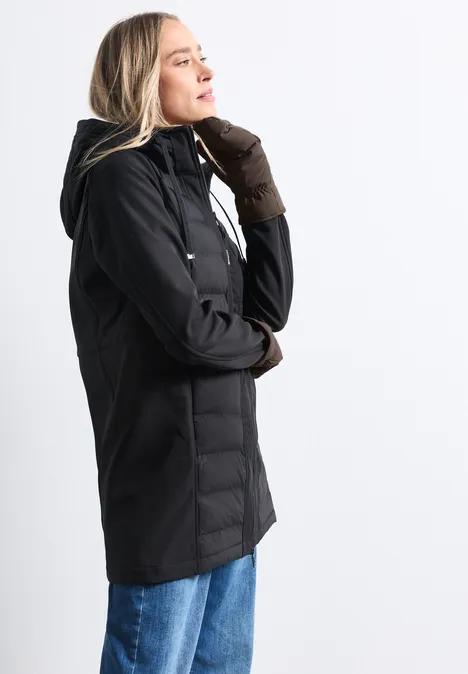 Frau im Profil trägt schwarze Jacke mit Kapuze, braune Handschuhe und blaue Jeans, vor hellem Hintergrund.