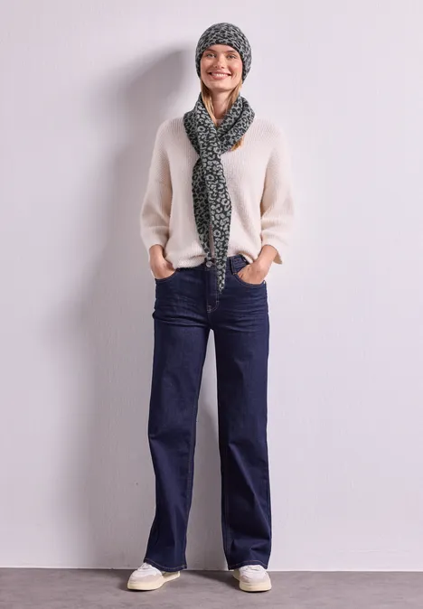 Eine lächelnde Person trägt eine graue Strickmütze, Leoparden-Schal, hellen Pullover und dunkle Jeans.