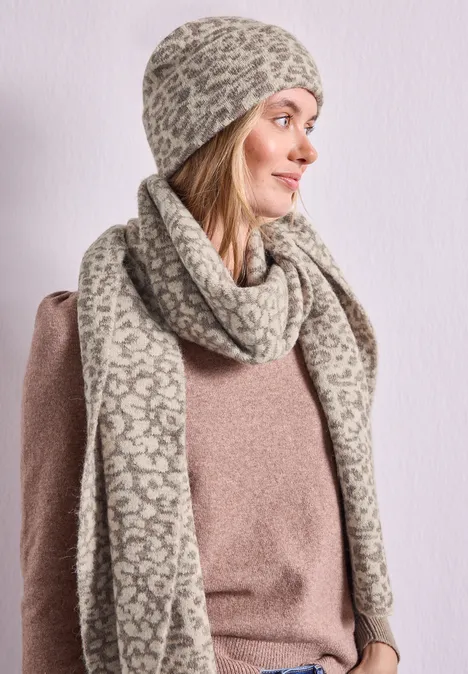 Frau mit beiger Mütze und passendem Schal mit animalischem Muster, trägt braunen Pullover.