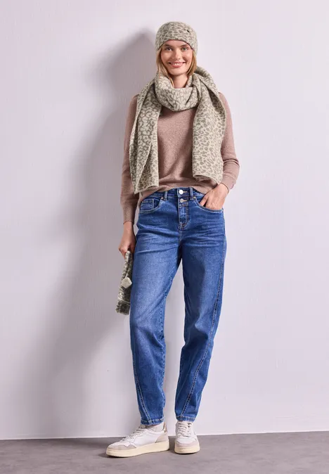 Frau im lässigen Look mit beiger Strickmütze und Schal, braunem Pullover, blauen Jeans und weißen Sneakers.