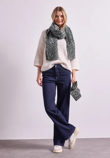 Frau in cremefarbenem Pullover, blauen Jeans, beigen Sneakers und grünem Strickschal mit passender Tasche.