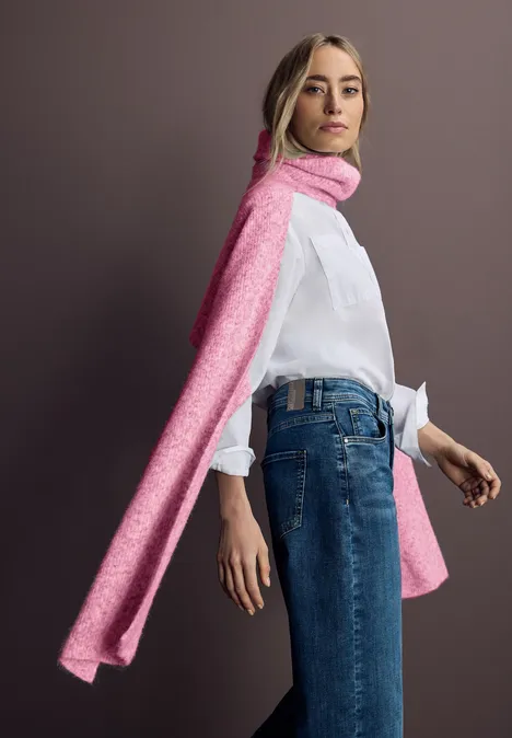Femme en chemise blanche, jean et longue écharpe rose debout devant un fond uni.