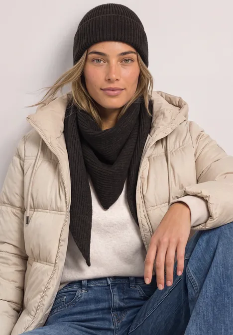 Beanie in Melange-Optik Black Junge Frau in beigefarbener Steppjacke, schwarzem Dreieckstuch und Mütze vor hellem Hintergrund.
