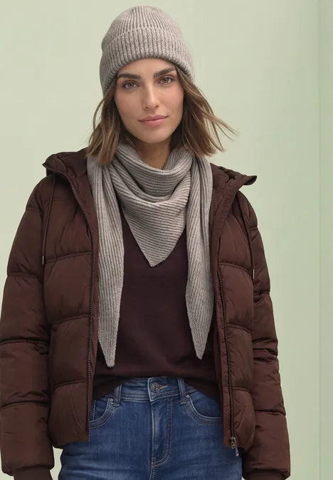 Frau in brauner Steppjacke, Jeans, grauem Schal und Mütze vor hellem Hintergrund.