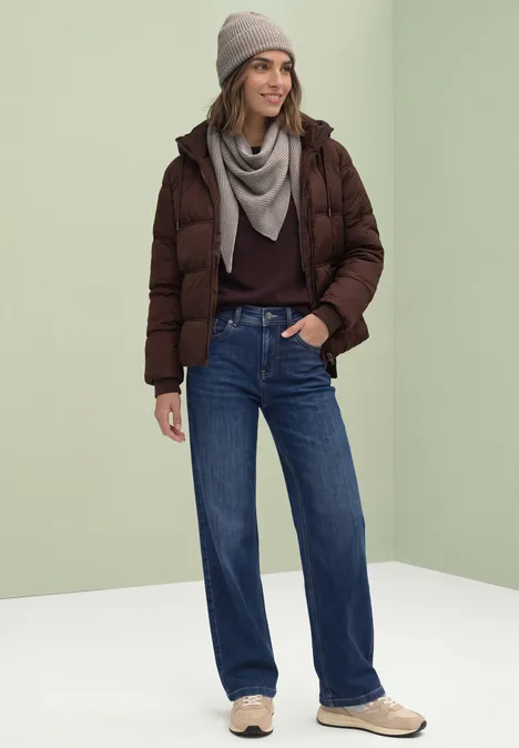 Frau in brauner Steppjacke, grauem Schal, Jeans und Mütze vor hellem Hintergrund.