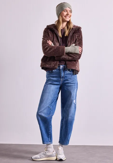Armwärmer in Melange-Optik Muddy Mocha Eine lächelnde Frau in Wintermode mit brauner Teddyjacke, Mütze, Jeans und weißen Sneakern.