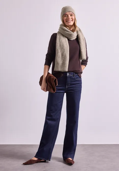 Eine lächelnde Frau in Mütze, Schal und Jeans, hält eine braune Tasche vor hellem Hintergrund.