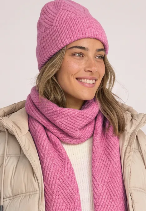 Frau mit pinker Strickmütze und passendem Schal, beiger Daunenjacke und freundlichem Lächeln.