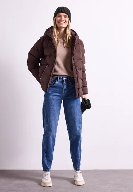 Eine junge Frau in brauner Steppjacke, beigem Pullover, blauen Jeans, weißen Sneakern und schwarzer Mütze lächelt.