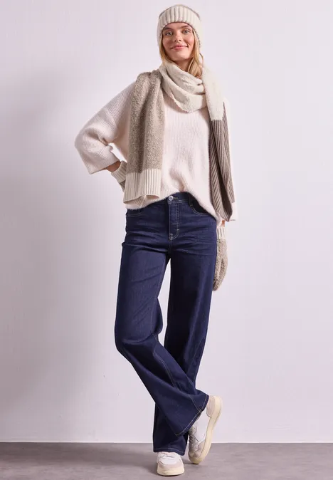 Eine Frau trägt einen Strickpullover, Schal und Mütze in Beige kombiniert mit weiter Jeans.