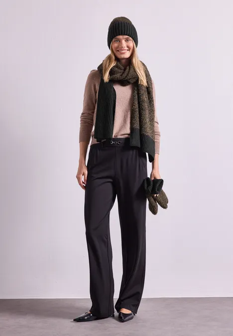 Bouclé-Mix Schal Black Frau in weiter schwarzer Hose, beigem Pullover, grünem Schal, Mütze und Handschuhen, lächelnd vor weißem Hintergrund.