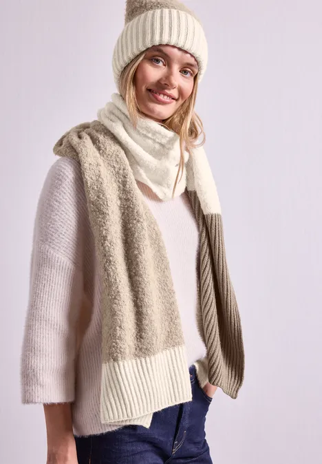 Bouclé-Mix Schal Muddy Mocha Eine Frau in cremefarbener Strickmütze, Schal und Pullover lächelt und trägt Jeans vor hellem Hintergrund.