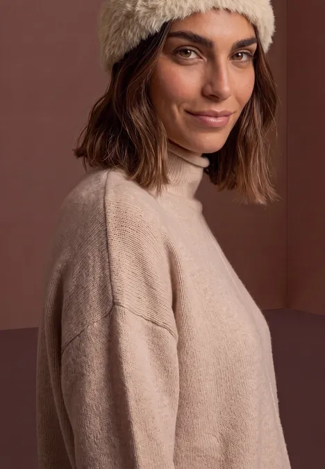 Stirnband aus Fake Fur smooth stone sand Frau mit braunem Bob, hellbeigem Strickpullover und Mütze, schaut lächelnd über die Schulter vor braunem Hintergrund.