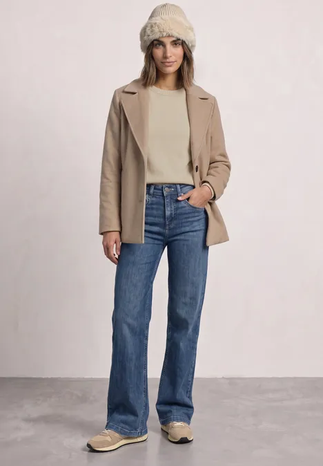Vrouw in crème trui, beige jas, blauwe bootcut jeans en een gebreide muts.