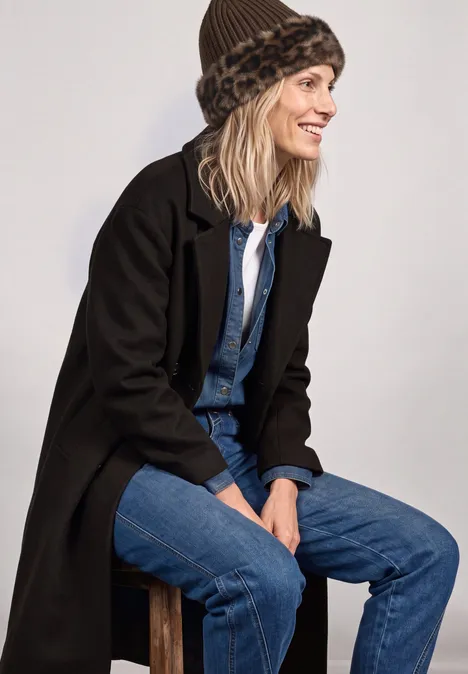Frau sitzt lächelnd seitlich auf einem Hocker, trägt schwarzen Mantel, Denim-Look und Leo-Fellmütze.