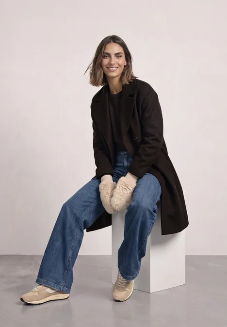 Eine lächelnde Frau sitzt auf einem weißen Block, trägt Jeans, Mantel, Fellhandschuhe und Sneaker.