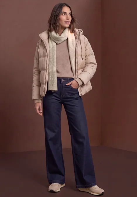 Frau trägt beige Steppjacke, Schal, Pullover und weite dunkelblaue Jeans vor braunem Hintergrund.