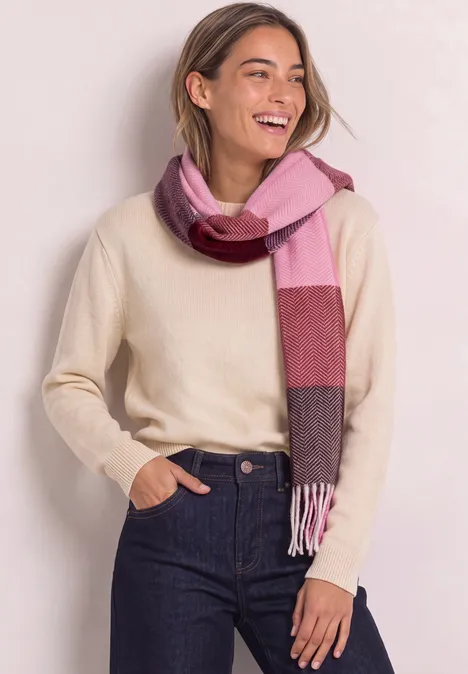 Eine lächelnde Frau trägt einen cremefarbenen Pullover, dunkle Jeans und einen rosa-braunen Schal.