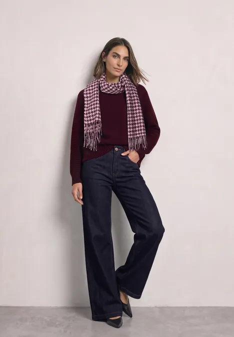 Frau in weinrotem Pullover, weitem Jeans, Hahnentritt-Schal und schwarzen Pumps vor hellem Hintergrund.