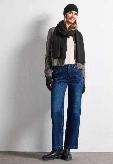 Vrouw in winteroutfit met muts, sjaal, handschoenen, jas en rechte jeans leunt glimlachend tegen muur.