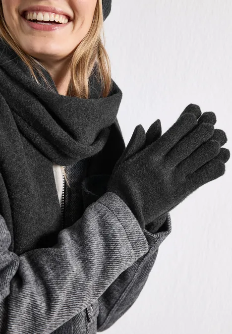 Lachend persoon draagt grijze sjaal, grijze jas en zwarte handschoenen voor een witte achtergrond.