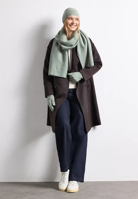 Gants en maille fine unie eucalyptus green mel. Frau trägt graue Mütze, Schal und Handschuhe, schwarzen Mantel, blaue Jeans und weiße Sneaker, lächelnd.