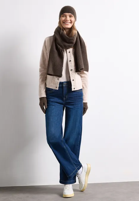 Feinstrick Handschuhe in Unifarbe auburn brown mel. Eine Frau mit braunem Schal, Handschuhen, beiger Jacke und blauen Jeans steht vor weißem Hintergrund und lächelt.