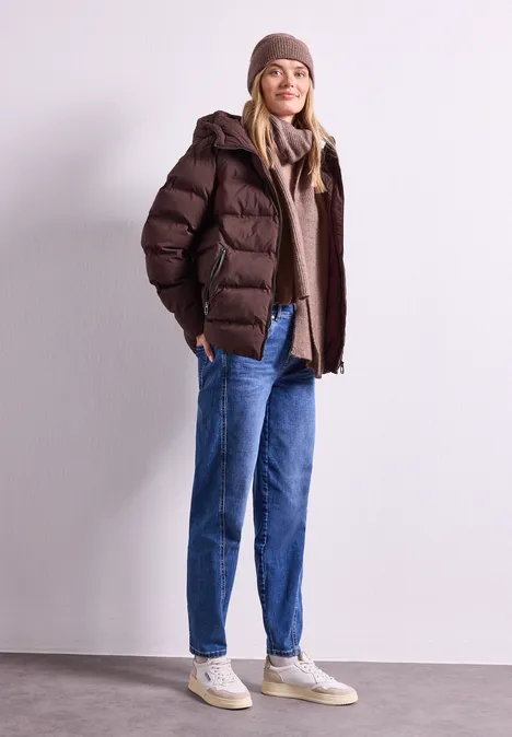 Beanie im Melange-Look vintage blush mel. Junge Frau trägt braune Steppjacke, Mütze, Jeans und Sneakers, lächelt vor schlichtem Hintergrund.