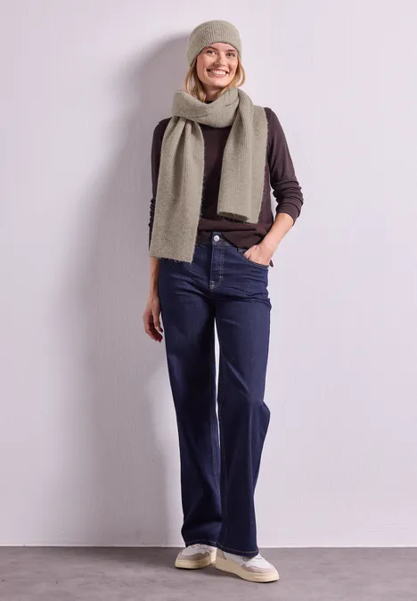 Beanie im Melange-Look barley beige mel. Frau in beiger Mütze und Schal, braunem Pullover, weiten Jeans und beigen Schuhen lehnt lächelnd an heller Wand.