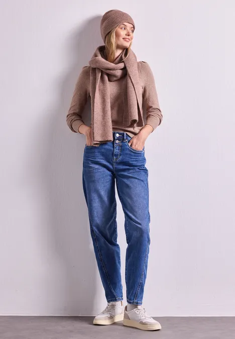 Schal im Melange-Look vintage blush mel. Junge Frau lehnt an weißer Wand, trägt Jeans, beigen Pullover, Schal, Mütze und Sneakers.