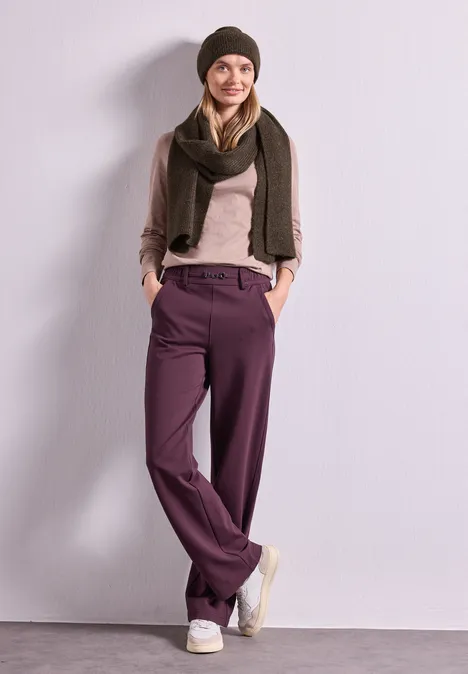 Schal im Melange-Look auburn brown mel. Eine Frau lehnt lächelnd an einer Wand und trägt lila Hose, beigen Pullover, braune Jacke, Schal und Mütze.