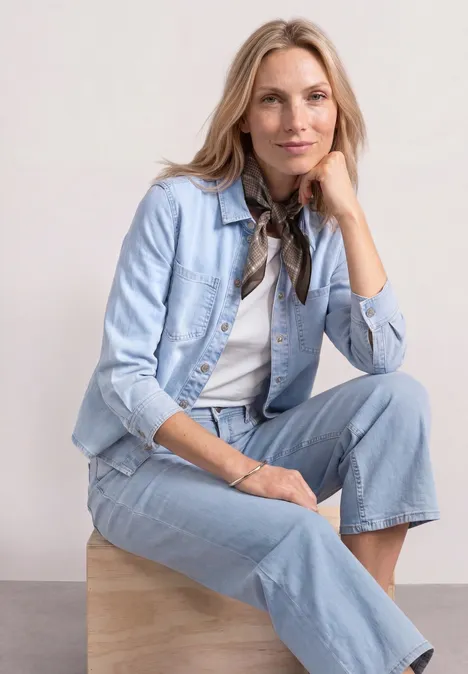 Bandana w kratkę silk brown Kobieta siedzi na drewnianym sześcianie, ma na sobie jasny jeansowy żakiet i spodnie z denimu.
