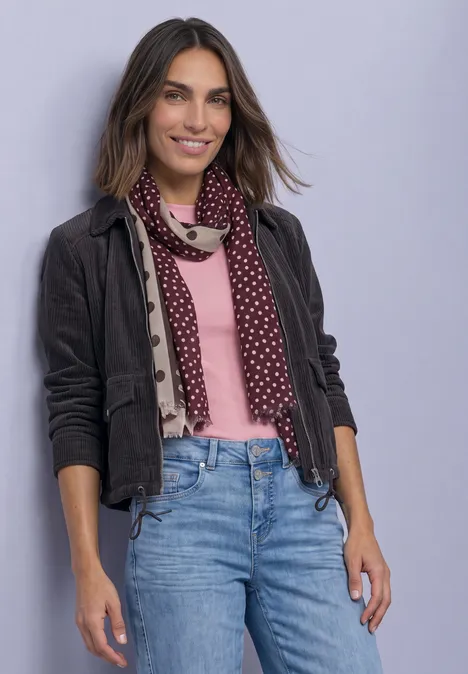 Eine lächelnde Frau mit braunen Haaren, rosa Pullover, grauer Jacke, gepunktetem Schal und Jeans vor hellem Hintergrund.