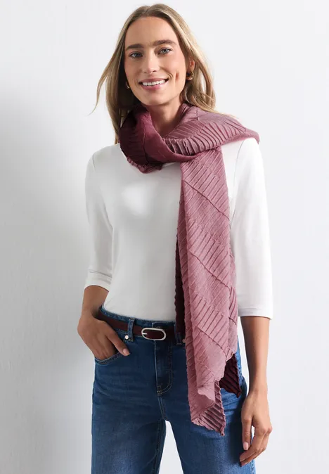Eine lächelnde Frau in weißem Shirt, Jeans und weinrotem Strickschal vor hellem Hintergrund.