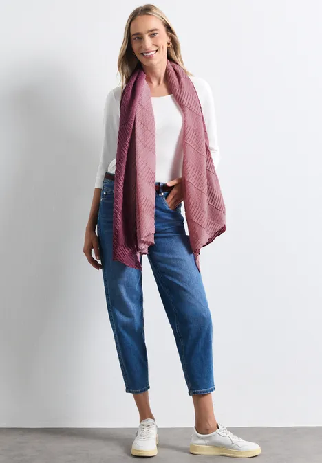 Frau lächelnd in weißem Shirt, blauen Jeans und rosa Schal vor weißem Hintergrund.