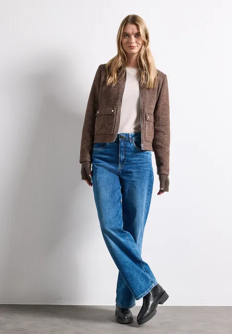 Armwärmer im Melange-Look auburn brown mel. Eine Frau in brauner Jacke, beigem Shirt, blauen Jeans und schwarzen Stiefeln steht vor weißem Hintergrund.