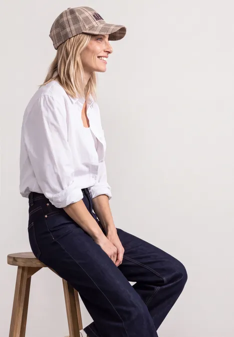 Frau mit weißer Bluse, Jeans und kariertem Cap sitzt lächelnd seitlich auf einem Hocker vor neutralem Hintergrund.