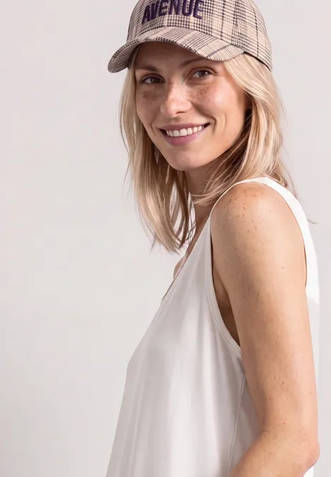 Blonde Frau mit Baseballcap, weißem ärmellosem Top, lächelt seitlich in die Kamera.