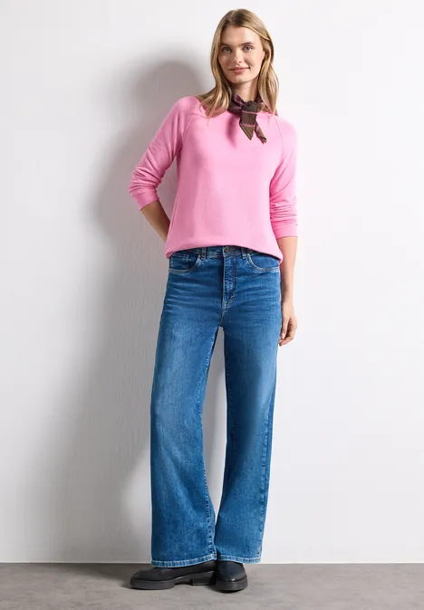Vrouw in roze trui, wijdvallende jeans en zwarte schoenen voor een witte, effen achtergrond.