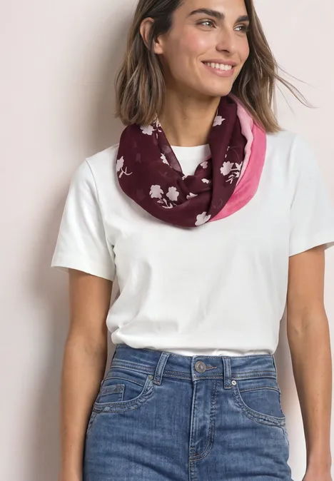 Loop-sjaal met bloemmotief rustic rouge Vrouw in wit T-shirt en spijkerbroek draagt een roze-bordeauxkleurige sjaal met witte bloemenprints.