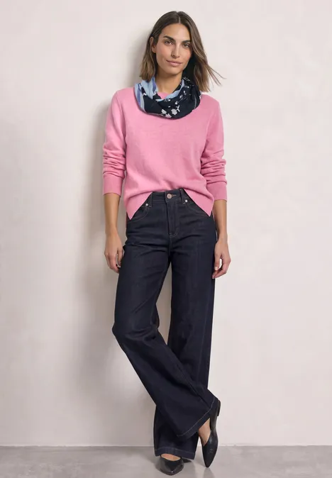 Model in roze trui, wijde donkere jeans, blauwe sjaal en zwarte pumps voor een lichte achtergrond.