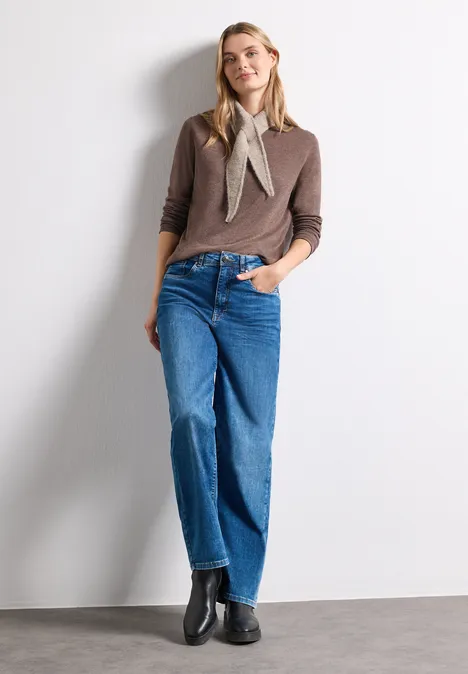 Mini-driehoekssjaal in effen kleur sanded amber mel. Vrouw voor witte muur, draagt bruine trui, sjaaltje, wijde blauwe jeans en zwarte laarsjes.