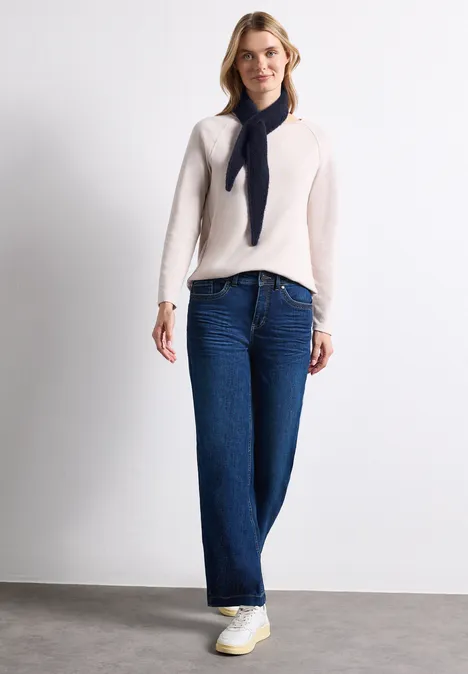 Mini-driehoekssjaal in effen kleur indigo night Vrouw met beige trui, brede jeans, witte sneakers en losjes geknoopt donkerblauw sjaaltje voor witte muur.