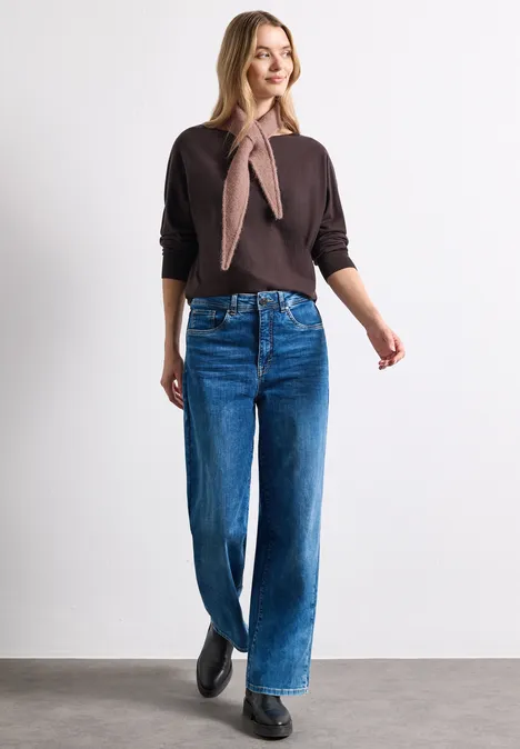 Frau in braunem Pullover, Halstuch, weiten Jeans und schwarzen Schuhen vor weißem Hintergrund.
