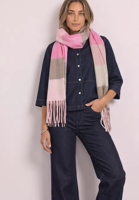 Eine Frau trägt einen dunkelblauen Denim-Overall und einen rosa-beigen Schal, sie steht vor hellem Hintergrund.