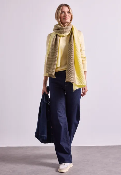 Une femme porte une écharpe jaune, un haut beige et un pantalon bleu foncé, tenant une veste en jean.